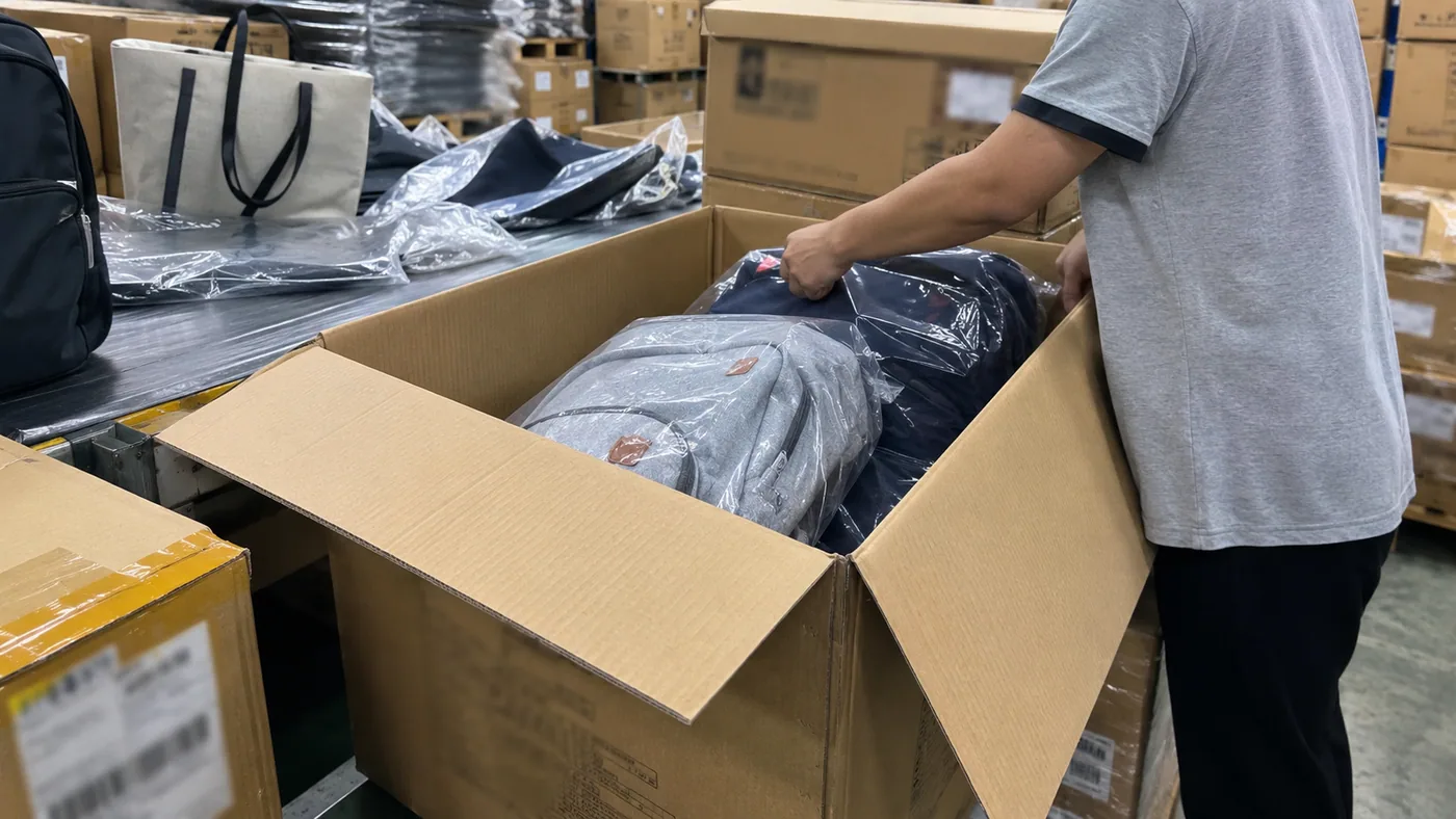 Smartphone-style factory packing area with backpacks and tote bags being packed into export cartons for North America