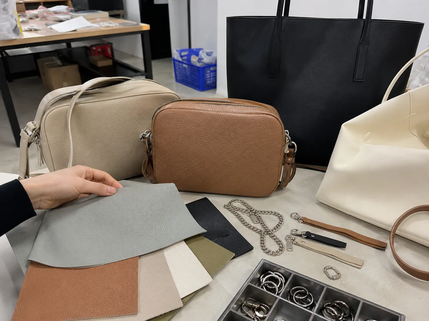 Smartphone-style sample-room table with fashion crossbody and tote bag samples, color swatches, chain strap, zipper pulls, and hardware tray