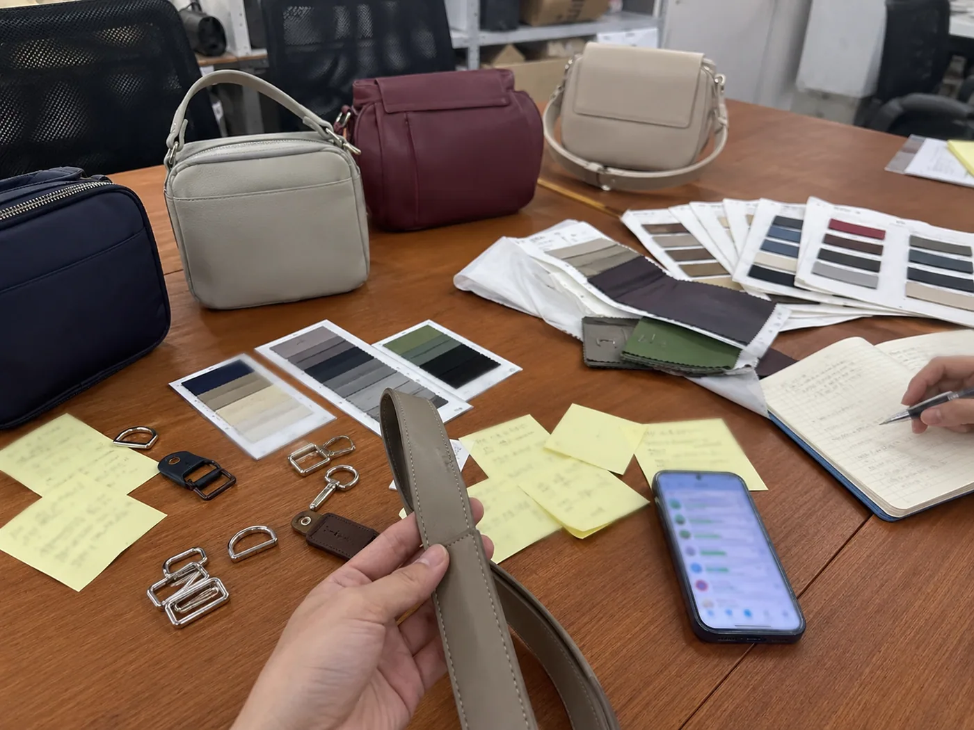 Smartphone-style Korean sample discussion table with fashion bag prototypes, color cards, hardware pieces, and blurred chat messages