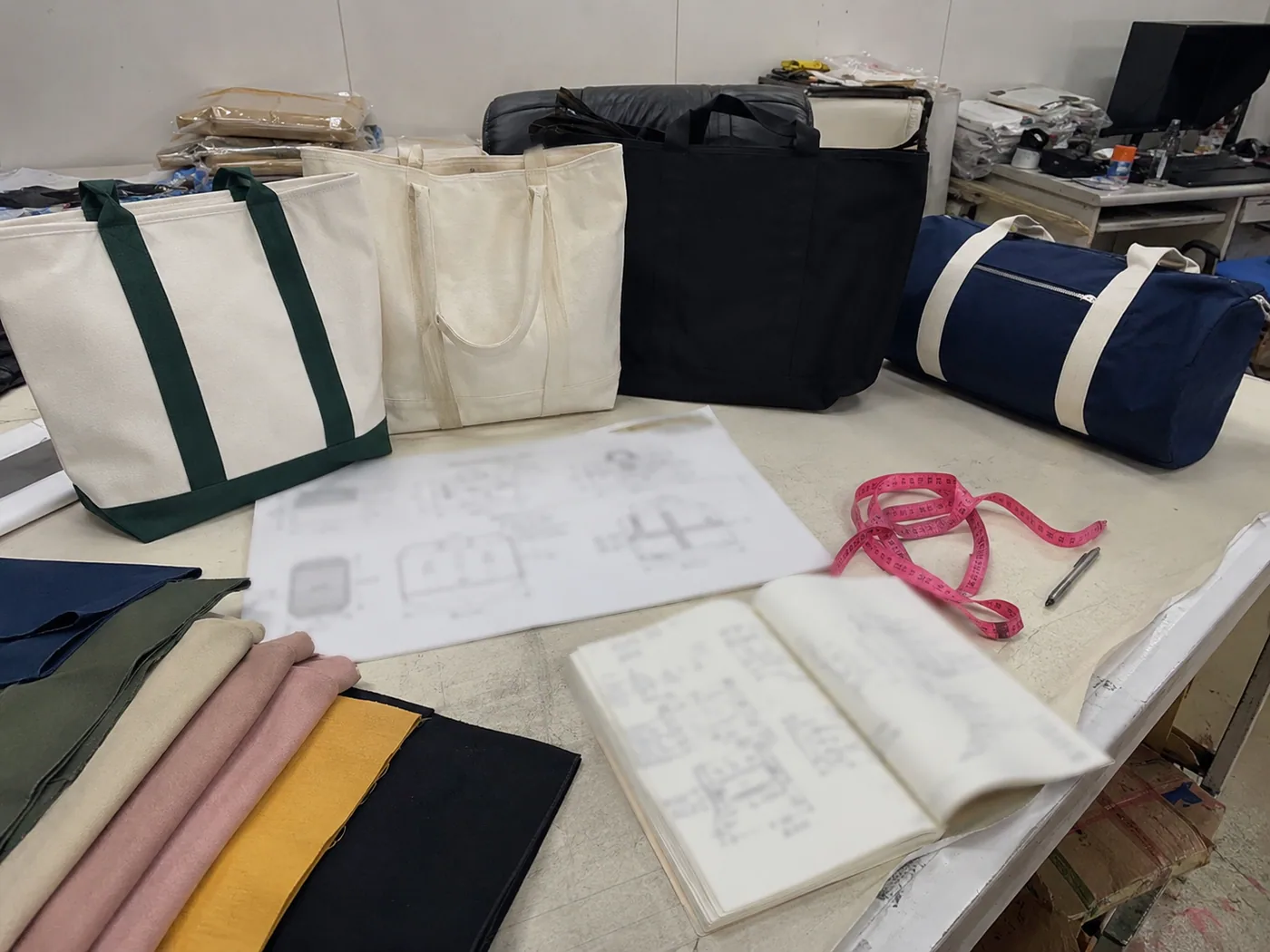 Smartphone-style Portuguese order planning table with tote samples, duffel sample, blurred carton sketch, fabric rolls, and measuring tape